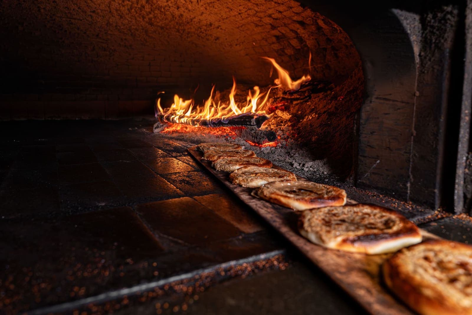 Flatbreads on a long peel inside a brick oven, flames along the far wall