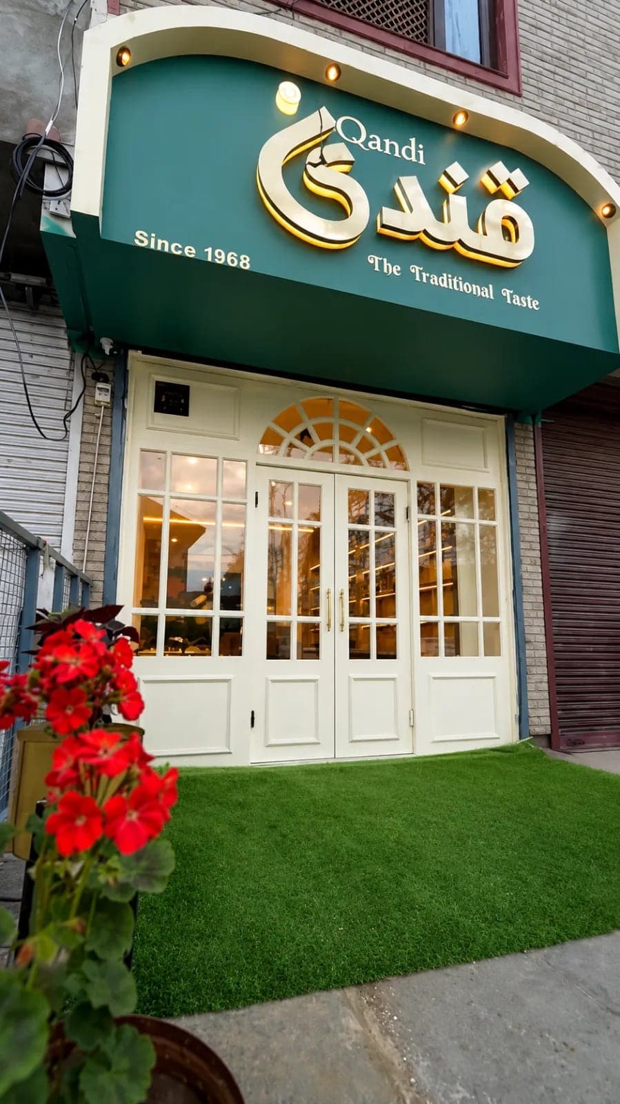 The Qandi Bakery shopfront on Janglat Mandi: a green awning with the name in gold Urdu lettering above 'Qandi', 'Since 1968' and 'The Traditional Taste', over white double doors with a fanlight, lit warm from inside